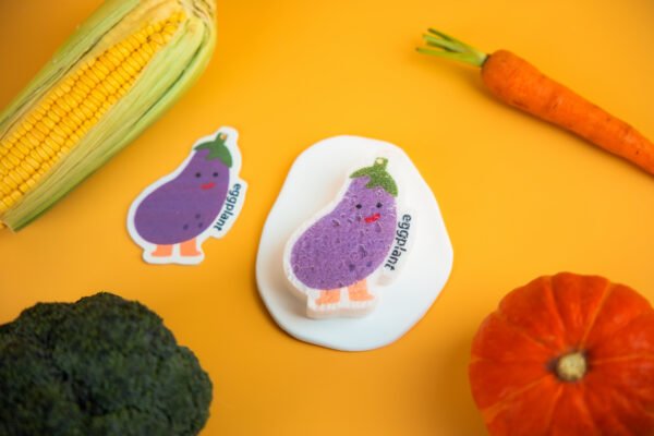 Vegetable Friends â Eggplant Biodegradable Sponge | Doyourclean Purple eggplant-shaped Doyourclean cellulose sponge on yellow backdrop with carrot and pumpkin decor.