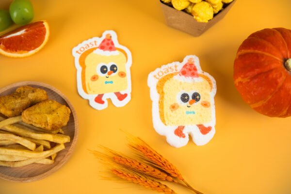 Tasty Friends â Toast Sponge | Doyourclean Eco Collection Two toast-shaped Doyourclean cellulose sponges with smiling faces, surrounded by snacks on yellow background.