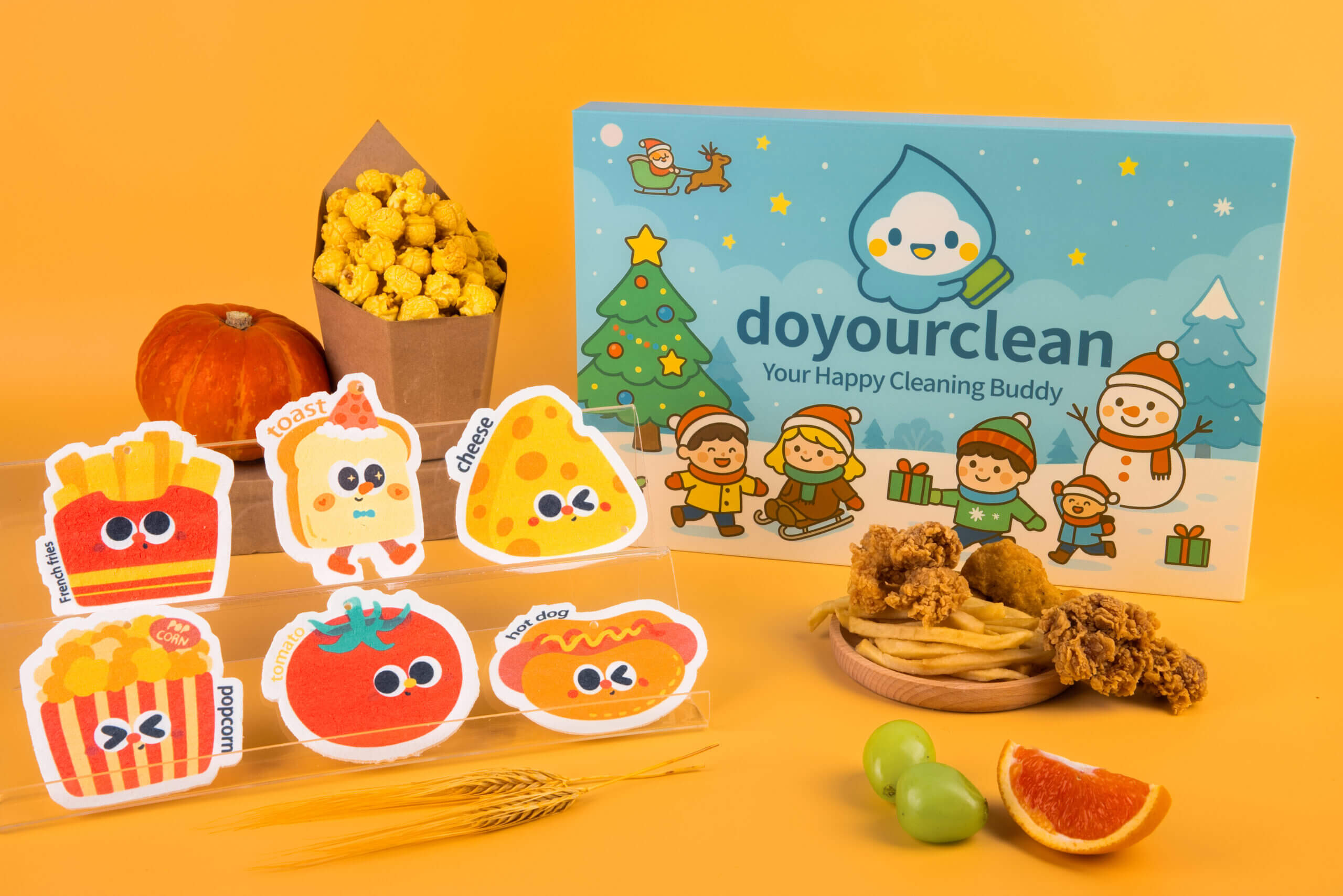 Doyourclean’s Tasty Friends cellulose sponge collection displayed with fries, toast, cheese, popcorn, tomato, and hot dog designs beside the gift box, surrounded by colorful snacks and props.